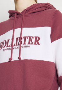 Primer plano de una persona que lleva una sudadera color burdeos y blanca con "HOLLISTER SOUTHERN CALIFORNIA" bordado en el pecho.