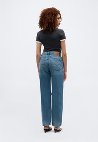 Γυναίκα με κατσαρά μαλλιά που φοράει μαύρο μπλουζάκι, τζιν ψηλόμεσο μπλε και μαύρα ψηλοτάκουνα σανδάλια, στέκεται με την πλάτη προς τα εμπρός σε ένα λευκό φόντο.