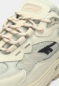 Šviesiai mėlynas sportbačio modelis iš tinklinės medžiagos ir odos, turintis kreminius raištelius su raudonomis detalėmis, tekstūruotas paviršius bei tamsiai mėlyną logotipo elementą.