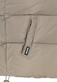 Beiger Puffermantel-Bereich mit gestepptem Textil, ausgestattet mit einer Seitentasche und einem Etikett mit der Aufschrift "SUBLEVEL" auf dem Stoff.