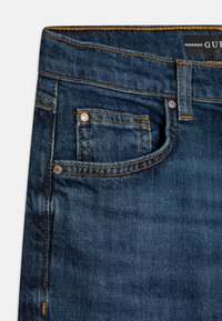 Mörkblå denimjeans med orange sömmar, som har en framficka och metallisk hårdvara. Tygets textur är slät med en lätt lyster.
