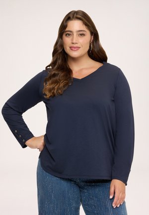Kvinde med bølget brunt hår iført øreringe med ringe, en navyblå langærmet V-hals top og blå jeans med glitrende lodrette striber.