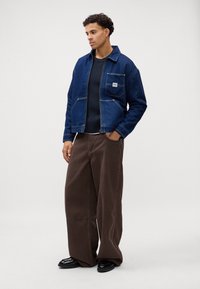 Donkerblauwe spijkerjack met een voorzak, gecombineerd met bruine wijde broek en zwarte schoenen. Soepele textuur, casual ontwerp.