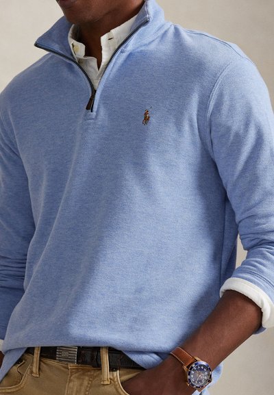 Homme portant un pull bleu clair à fermeture éclair quart sur une chemise blanche, pantalon beige, et une montre-bracelet en cuir marron avec une lunette bleu marine.