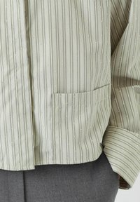 Lys grønstribet skjorte med metalliske detaljer. Har en lomme på venstre side og blødt stof med et struktureret mønster.