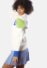 Cremfarbener Rippstrickpullover mit blauen und grünen Akzenten, der einen großen blauen Buchstaben zeigt. Kombiniert mit einem plissierten blau-rosa karierten Rock.