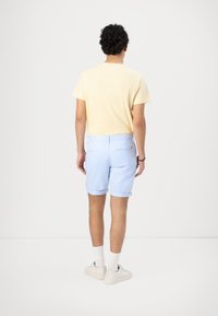 Camiseta amarilla clara, pantalones cortos azules claros enrollados, calcetines blancos y zapatillas beige. El atuendo es sencillo y casual, con materiales suaves.