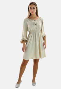 Cremefarbenes Kleid mit langen Ärmeln, geraffter Taille, vertikaler Struktur und drei Knöpfen am Ausschnitt. Silberne flache Schuhe mit Pailletten.