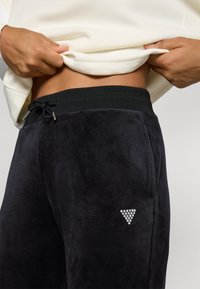 Zwarte joggingbroek van een zachte, suede-achtige stof met een trekkoord in de taille en een strass driehoeklogo op de voorzak.