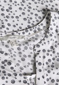 Vit blus med små svarta blommönster, rund halsringning och kort knappslå. Tyget är lätt och mjukt. Varumärkesetikett synlig.