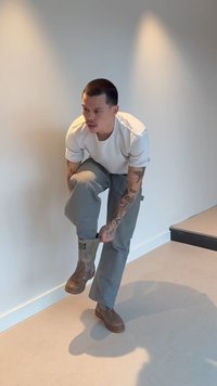 Homme tatoué portant une chemise blanche et un pantalon gris enfilant des bottes marron en position debout près d'un mur blanc et d'un escalier à l'intérieur.