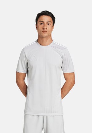 Mann steht mit den Händen hinter dem Rücken, trägt komplett weiße Adidas-Sportbekleidung und schaut nach vorne vor einem schlichten hellen Hintergrund.