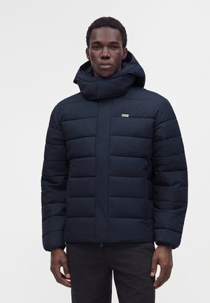 Homme portant une veste matelassée noire avec un col montant et une capuche, debout devant un fond clair uni.