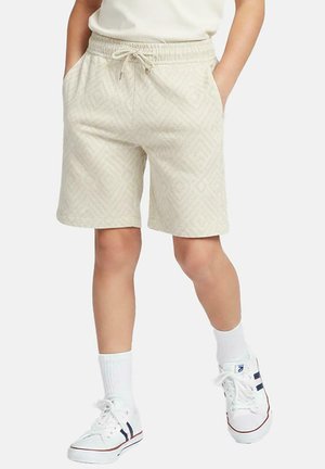 Persoon draagt beige, gestructureerde shorts met trekkoord, witte enkelsokken en witte sneakers met marineblauwe en rode strepen, staat met handen in de zakken.