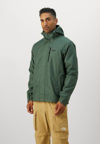Chaqueta impermeable verde con capucha, que cuenta con un bolsillo en el pecho con cremallera y un logo en la parte izquierda del pecho, combinada con pantalones cargo beige.