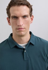 Camisa polo de color azul verdoso con cuello, con tapeta de botones y un pequeño logo. Textura suave, presentada sobre una persona con fondo neutro.