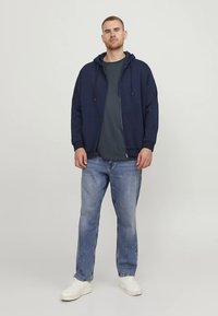 Sudadera con capucha azul marino con cordones, cremallera frontal y bolsillo tipo canguro, combinada con una camiseta gris y jeans azul claro. Zapatillas blancas completan el conjunto.