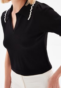 Black knitted top with a deep V-neck, featuring a white ruffled collar and short, puffed sleeves. Smooth texture, fitted design.