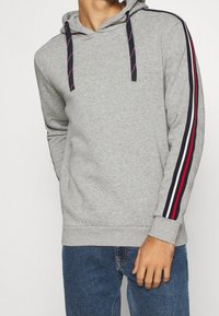 Haut à capuche gris avec cordon de serrage, présentant des rayures bleu marine et rouges le long des manches. Fabriqué en tissu doux, avec une coupe décontractée et des poignets côtelés.