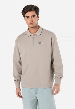 Beige langærmet polo-shirt med lynlås, der har en krave og et sort broderet logo på brystet, lavet af blødt bomuldsstof.