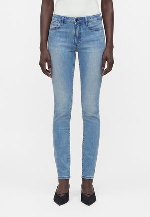 Γυναίκα που φοράει ανοιχτό μπλε τζιν στενής εφαρμογής, λευκό τοπ και μαύρες μυτερές γόβες, στέκεται μπροστά σε ένα απλό λευκό φόντο.