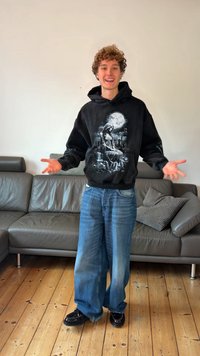 Črna jopa s kapuco z grafičnim potiskom sedeče figure, združena z ohlapnimi modrimi kavbojkami in črnimi čevlji, na lesenem tlaku blizu sivega kavča.