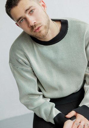 Pull rayé en gris clair et noir, col rond, poignets côtelés et tissu texturé. Le modèle montre des tatouages sur les mains, assis contre un fond neutre.