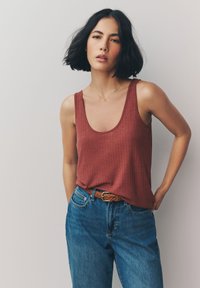 Débardeur à col rond de couleur rouille, à côtes, associé à un jean en denim bleu taille haute et une ceinture tressée marron.