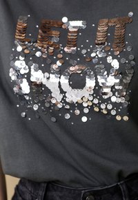 Grijze T-shirt met glinsterende tekst die "LET IT NOW" zegt in verschillende metallic kleuren, met een zachte katoenstructuur.
