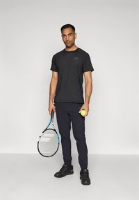 T-shirt desportiva preta de manga curta com logótipo, combinada com calças de desporto azul-marinho e ténis pretos. Segurando uma raquete de ténis e uma bola.