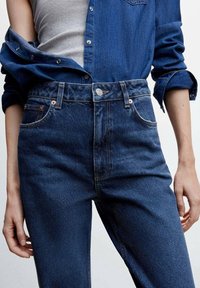 Mørkeblå denimjeans med høj talje, der har fem lommer og bronze hardware. Stoffet har en glat tekstur med diskrete slidser.