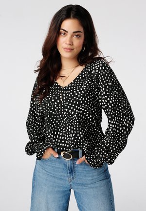 Femme aux longs cheveux foncés portant un chemisier à pois noir et blanc, un jean bleu et une ceinture noire avec une boucle dorée, debout les mains dans les poches.