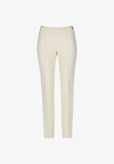 Gaiši bež bikses, kas izgatavotas no gludas auduma. Tām piemīt pieguļošs dizains ar priekšējiem šuviem un elastīgu jostu. Nav kabatu vai rakstu.