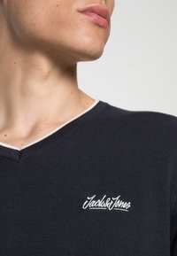 Zbliżenie na szyję i szczękę osoby noszącej czarny sweter z dekoltem w serek, wykończony białą lamówką oraz białe logo "Jack & Jones" na klatce piersiowej.