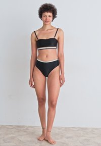 Črn bikini komplet z bandeau zgornjim delom in visokim pasom, ki ima bele črtaste detajle. Gladka tkanina, minimalističen dizajn.