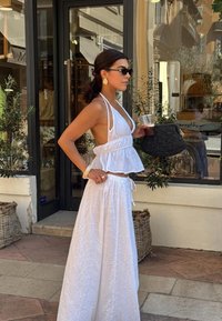 Weißes, ärmelloses Peplum-Top mit Spitzenakzenten, kombiniert mit einem langen weißen Rock mit floralen Stickereien. Die Accessoires umfassen eine schwarze Tasche.