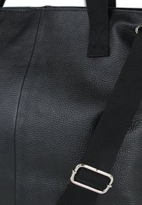 Zwarte leren totebag met een gestructureerd oppervlak, voorzien van stoffen handvatten en een verstelbare riem met een zilverkleurige metalen gesp. Geen zichtbare patronen of accenten.