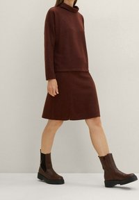 Hnědý pletený rolák v kombinaci s odpovídající sukní ve tvaru A ke kolenům. Modelka má na sobě hnědé kotníkové boty s širokou podrážkou.