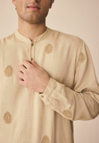 Hellbeige Kurta aus strukturiertem Stoff, mit goldener Stickerei in floralem Muster und einem geknöpften Rundhalsausschnitt sowie langen Ärmeln.