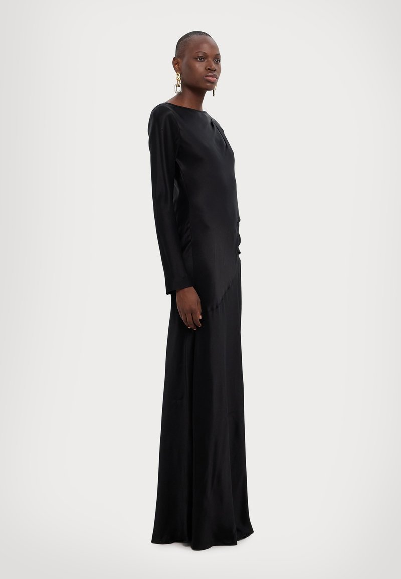 Vestido largo de satén negro con un corte ajustado, mangas largas y una silueta recta. Presenta un detalle drapeado en un lado y una textura suave.