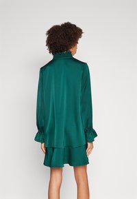Abito in satin verde con colletto arricciato e maniche a sbuffo; presenta un orlo a strati e una texture liscia. La vista mostra il retro.