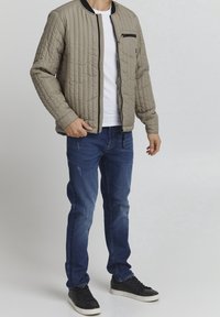 Veste bomber beige matelassée avec un col noir, comportant une poche zippée, associée à un jean bleu déchiré et des baskets noires.