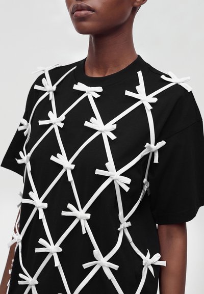 Chemise noire à manches courtes avec des rubans blancs croisés noués en nœuds couvrant le devant et les épaules, portée par une personne à la peau foncée.