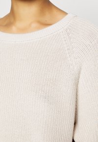 Pull tricoté beige clair avec un col large et des manches raglan. La texture est légère et respirante, avec un design simple.