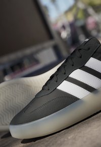 Scarpe sportive nere con strisce bianche e suola traslucida posizionate su una superficie di cemento alla luce naturale.
