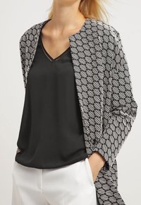 Top di seta nera con scollo a V, abbinato a una giacca aperta nera e bianca con motivo geometrico, indossata su pantaloni chiari.