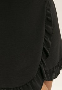 Jupe noire avec un ourlet volanté et des détails sur les côtés, réalisée en un tissu léger et doux avec une texture lisse.