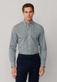 Camisa de botones a cuadros verdes y blancos, con cuello y mangas largas, combinada con pantalones azul marino oscuro. Presenta costuras cuidadas y un detalle de logo.