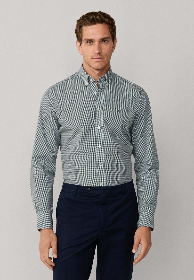 Camisa de botones a cuadros verdes y blancos, con cuello y mangas largas, combinada con pantalones azul marino oscuro. Presenta costuras cuidadas y un detalle de logo.