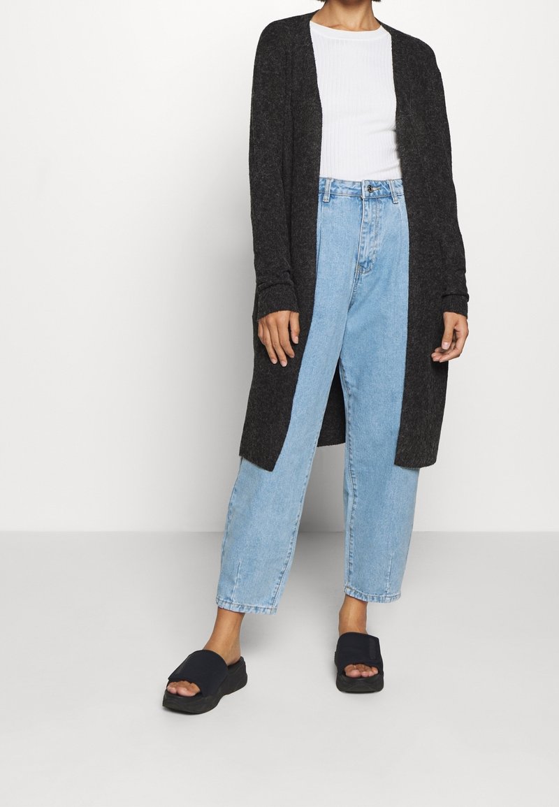 Lång mörk cardigan över en vit ribbad topp, kombinerad med högmidjade ljusblå jeans och svarta slidesandaler. Avslappnad design och passform.